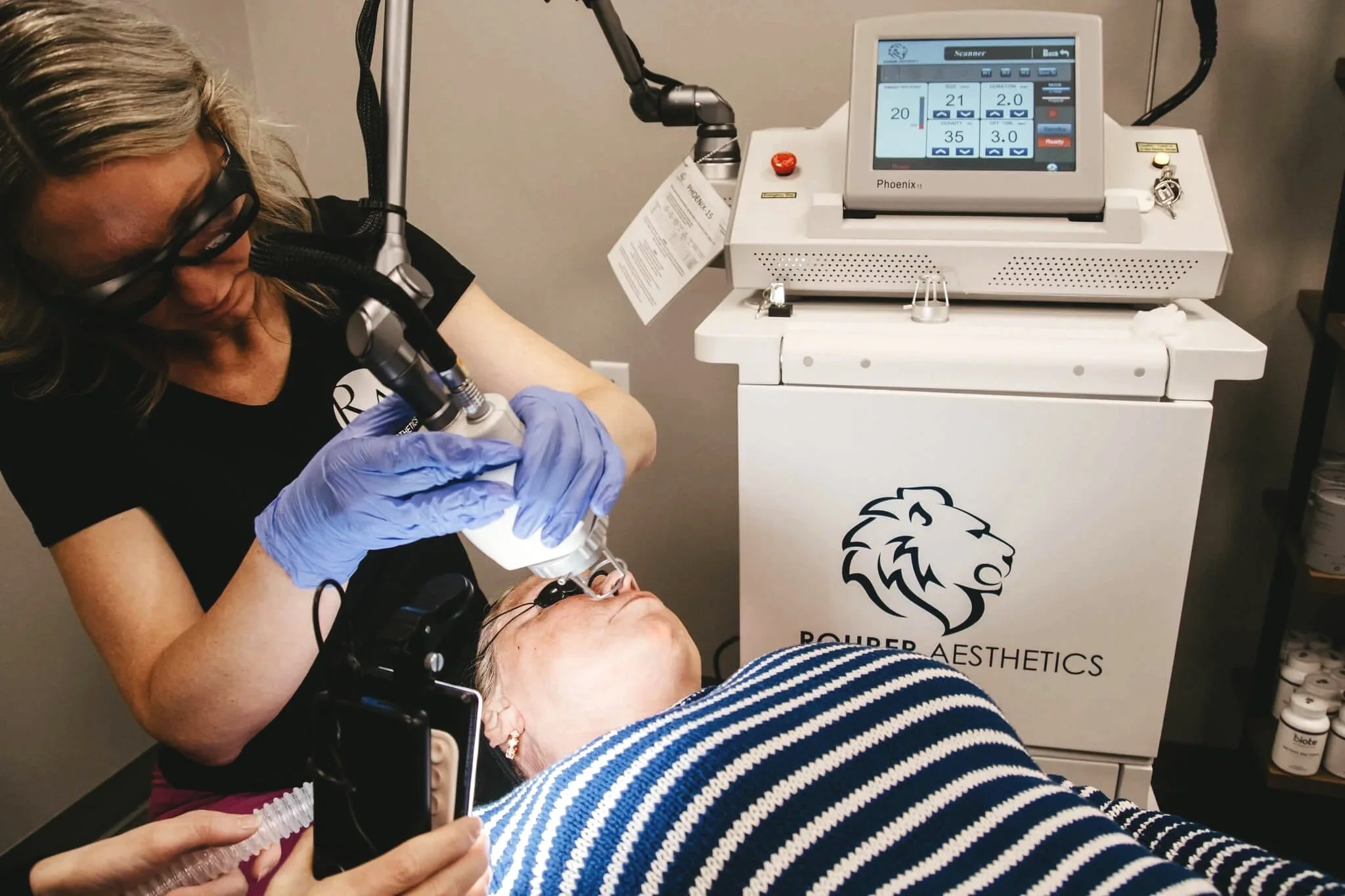 A woman receiving a cosmetic procedure on her face using a specialized machine operated by a technician wearing gloves. The technician is focusing on the woman's face with a microscope attached to the device. The procedure is taking place in a clinic with a machine labeled 'Pouper Aesthetics'.