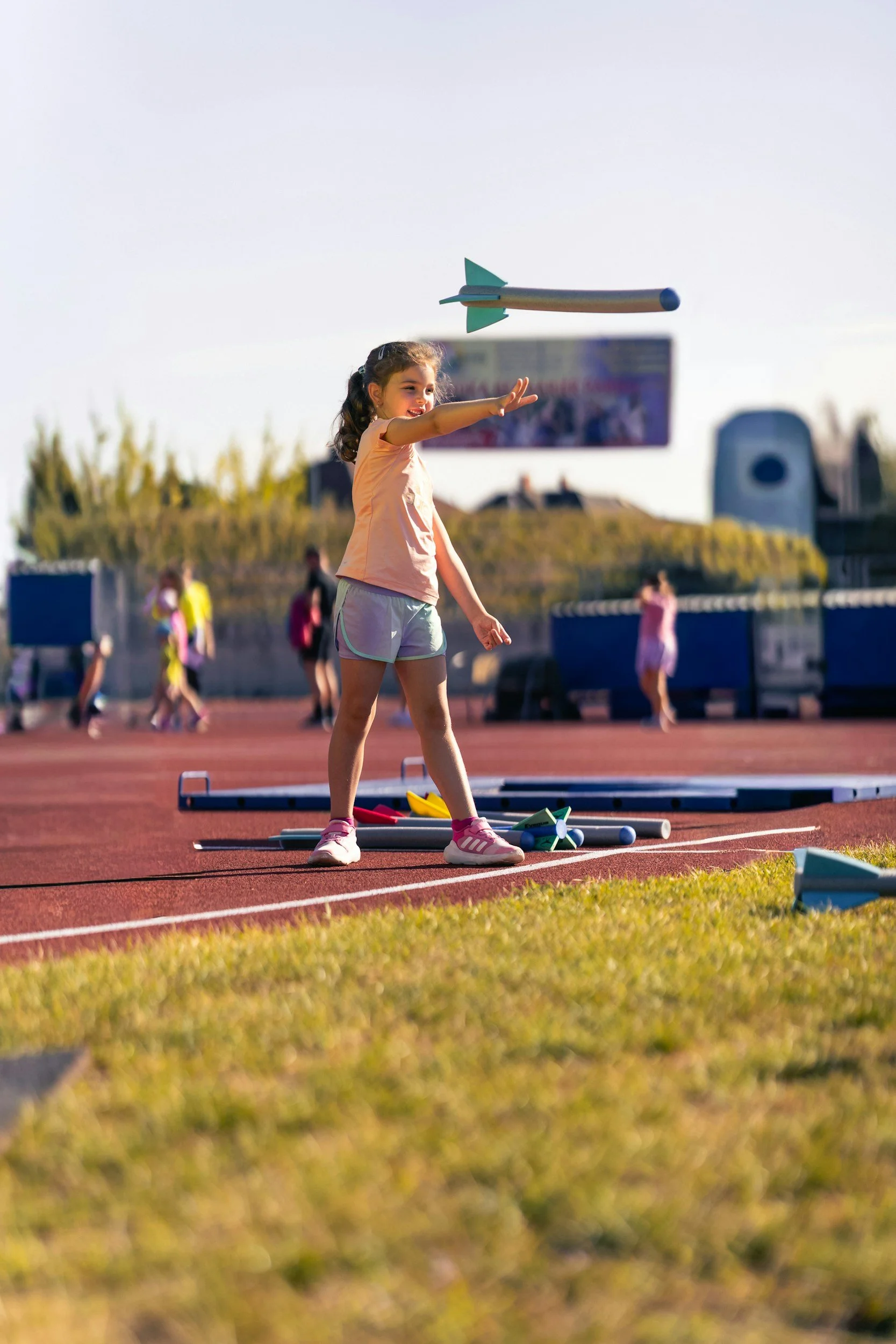 A young girl at an outdoor athletics track releasing a small model rocket into the air. She is smiling and reaching out with her right hand, with other children and objects visible in the background.