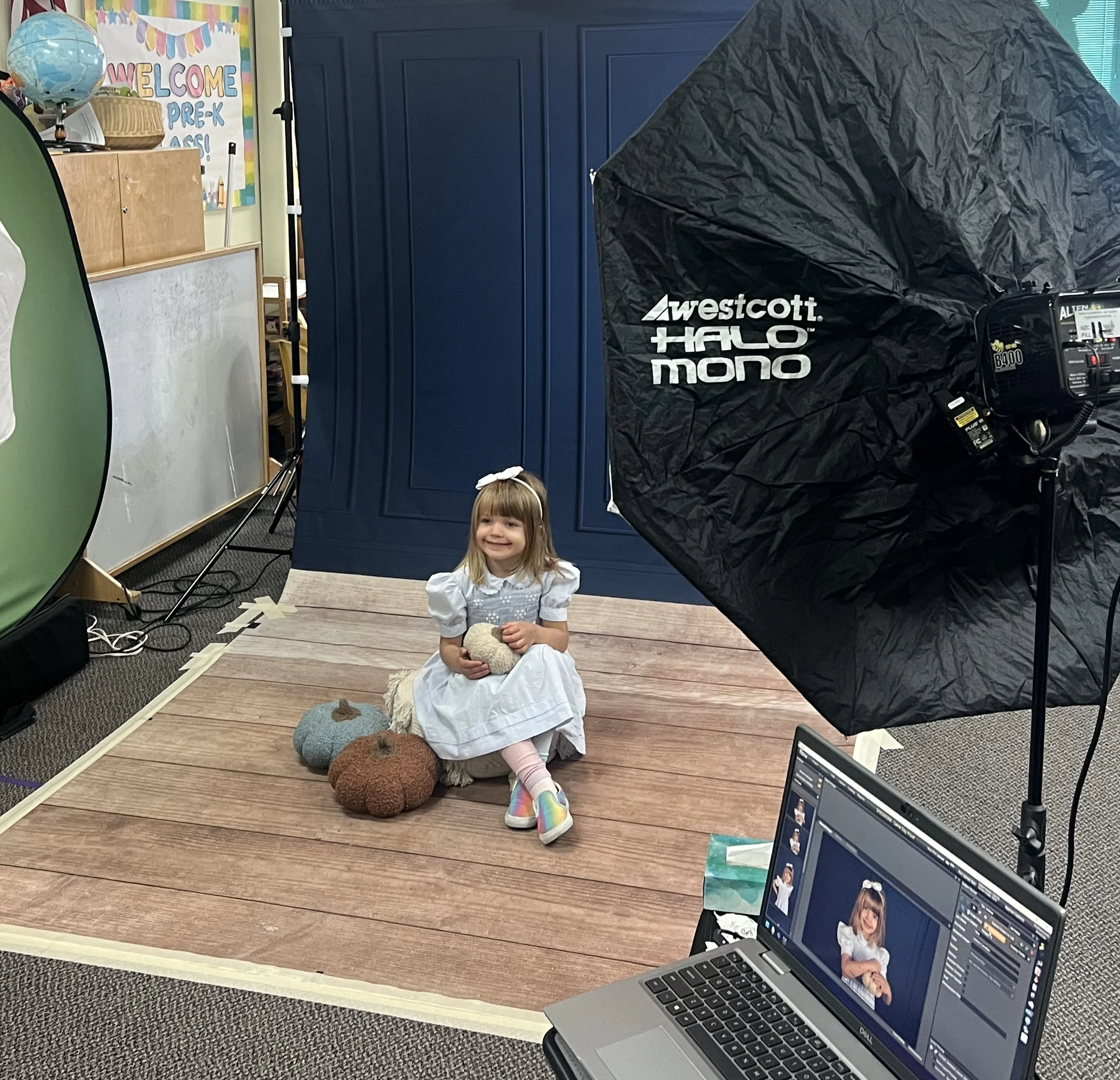A young girl in a white dress with rainbow shoes sitting on a wooden floor with plush pumpkins, holding a small doll or stuffed animal, in a photography studio setting with professional lighting equipment and monitor displaying her photo.
