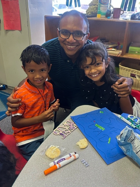 A man with glasses smiling and hugging two children, a boy in an orange striped shirt and a girl in a black shirt, sitting at a table with art supplies and a blue poster.