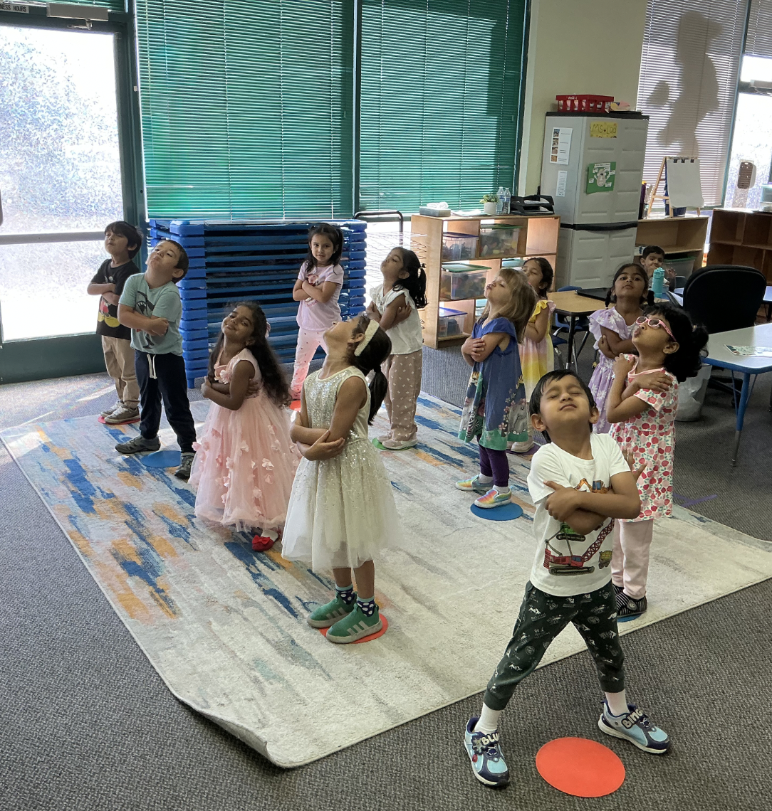 Children in a classroom standing on colorful circles, with some wearing costumes, participating in an activity.