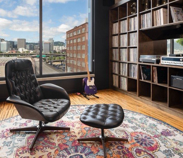 Modern office breakout area with vintage lounge chair and ottoman on a large round rug, floor-to-ceiling custom bookshelves styled with art, memorabilia, and books, and expansive city views of Portland through full-height windows