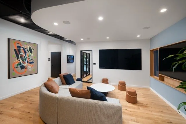 Creative office breakout lounge featuring a large curved gray sofa, contemporary artwork, architectural lighting, and a multimedia screen designed for team collaboration and presentations