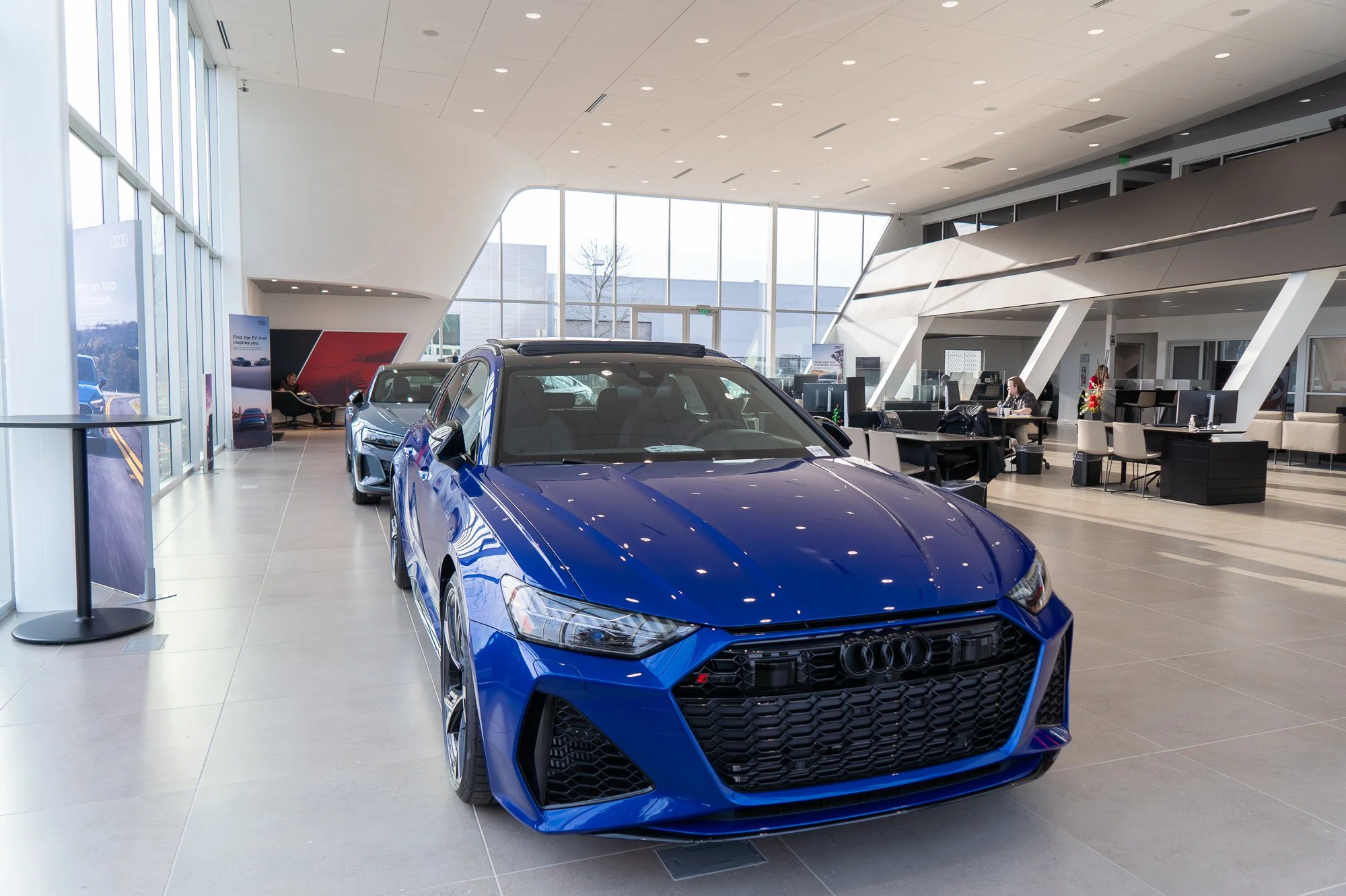 A blue Audi sports car displayed in a modern car dealership showroom with floor-to-ceiling windows and several employees working at desks in the background in Wilsonville, Oregon