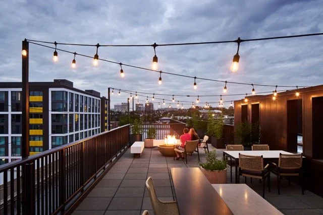 Rooftop patio on a multi-family building in Portland, Oregon featuring lounge seating, a fire pit, and string lights glowing beneath early evening clouds