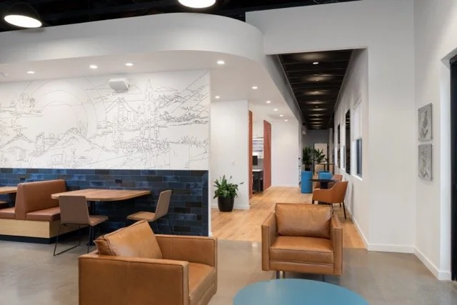 Employee kitchen lounge in a commercial office featuring brown leather club chairs, a custom hand-painted mural above built-in banquette seating, and a warm, modern atmosphere