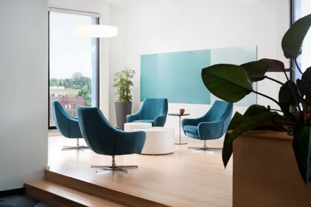 Modern office lounge in Portland, Oregon featuring blue armchairs, a white coffee table, biophilic design elements, and a large window with expansive views