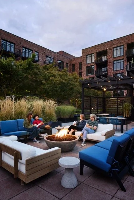 Rooftop lounge seating with people gathered around a modern gas fire pit at a multi-family residential property in Portland, Oregon, featuring outdoor sofas and lush landscaping