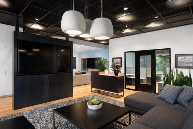 Modern office lounge area in Hillsboro, Oregon featuring black and white decor, a gray sectional sofa, wall-mounted TV, oversized white pendant lights, black perforated mesh drop ceiling, and white oak wood floors