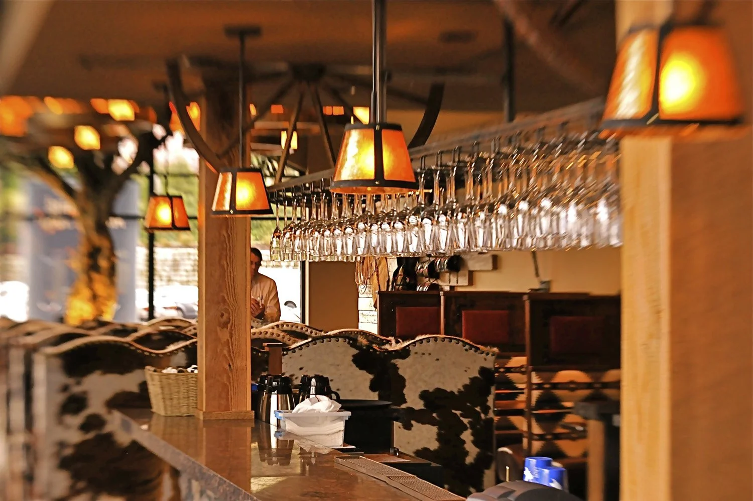 Interior of a restaurant or bar with hanging glasses and warm orange lighting, cowhide bar stools, and a bartender in the background.