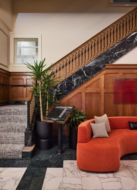 Historic commercial office building lobby in Downtown Portland, Oregon featuring a grand marble and wood staircase, modern burnt orange sofa, large potted plant, marble floors, and contemporary acrylic artwork