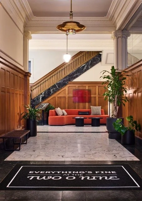 Historic lobby in downtown Portland featuring a grand staircase, antique glass chandelier, detailed wood wainscoting, marble floors, and a modern seating area with a custom orange sofa, potted plants, and contemporary artwork