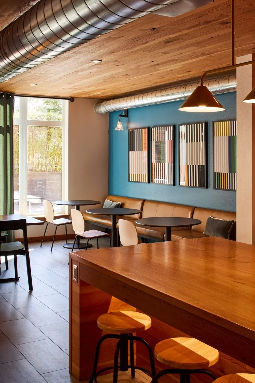 Residential community space in a multifamily building in Portland, Oregon featuring a warm wood ceiling, kitchen island, bright blue accent wall with modern art, and under-counter lighting illuminating contemporary counter stools