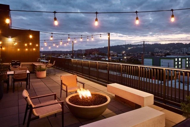Rooftop terrace of a multifamily commercial building in Portland, Oregon, featuring string lights, outdoor lounge seating, a modern fire pit, and panoramic city views at dusk