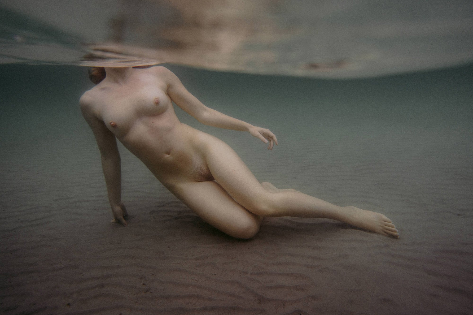  An underwater shot of a womens naked body rested on the sea bed, her head not visible as it’s above the water. 
