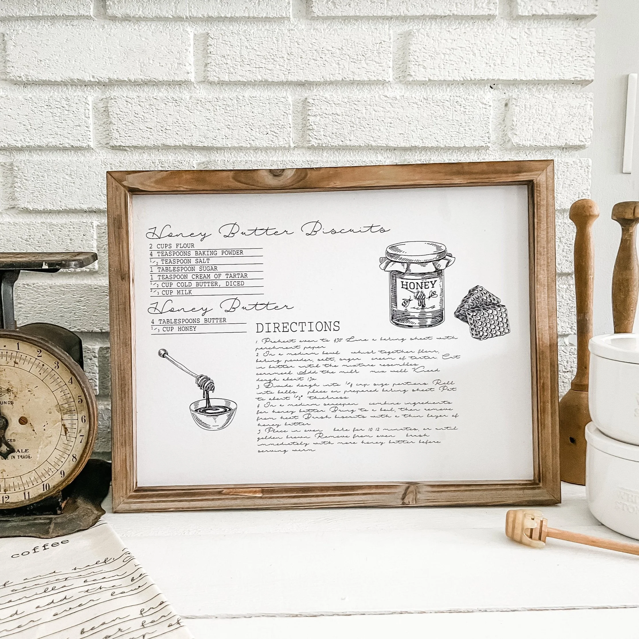 Framed recipe for honey butter biscuits with hand-drawn illustrations of honey jar and honeycomb, on a white countertop with vintage scale, honey dipper, and white ceramic containers nearby.