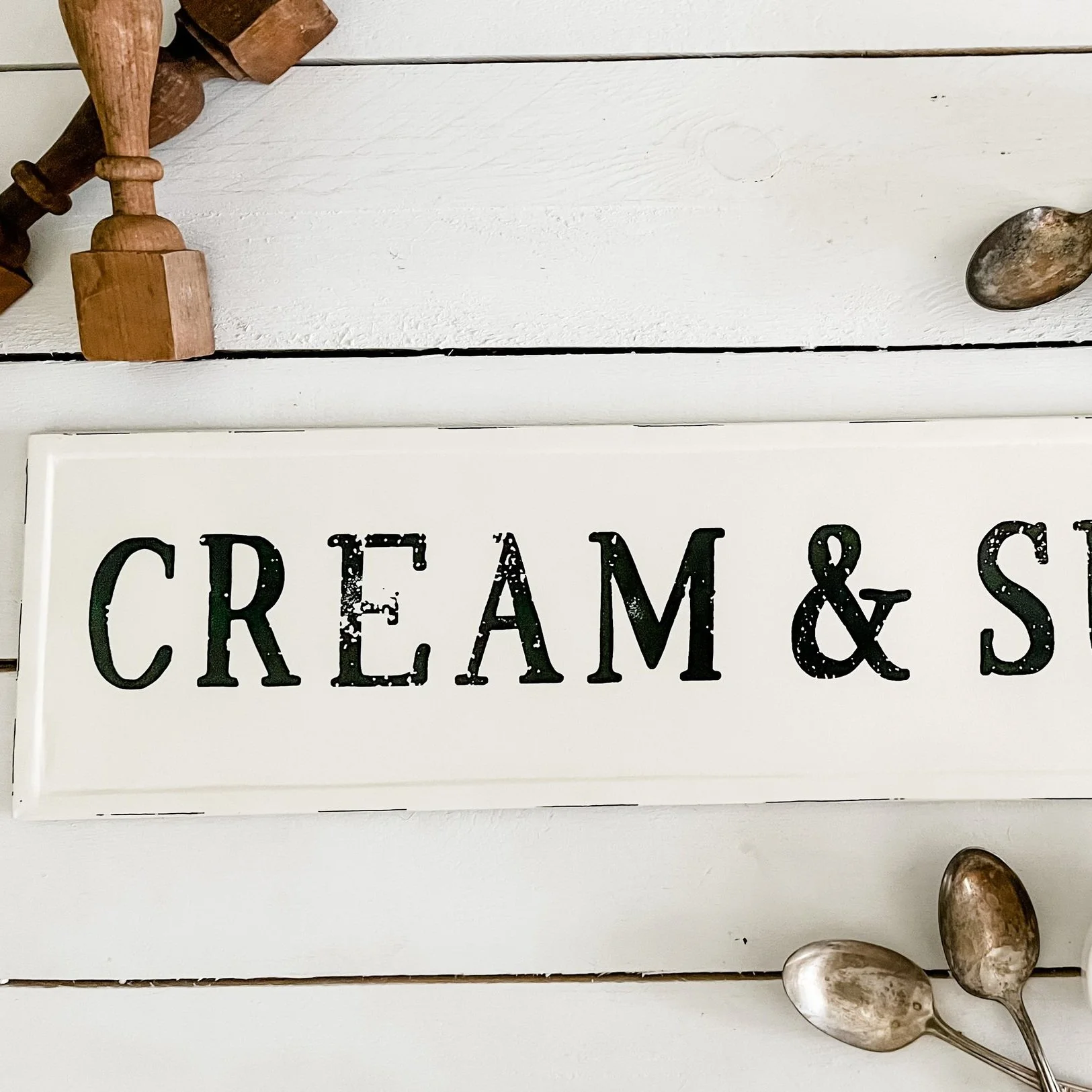 Close-up of a white sign with black distressed letter text that reads 'CREAM & S...' on a white wooden surface, with antique spoons and wooden utensils around it.