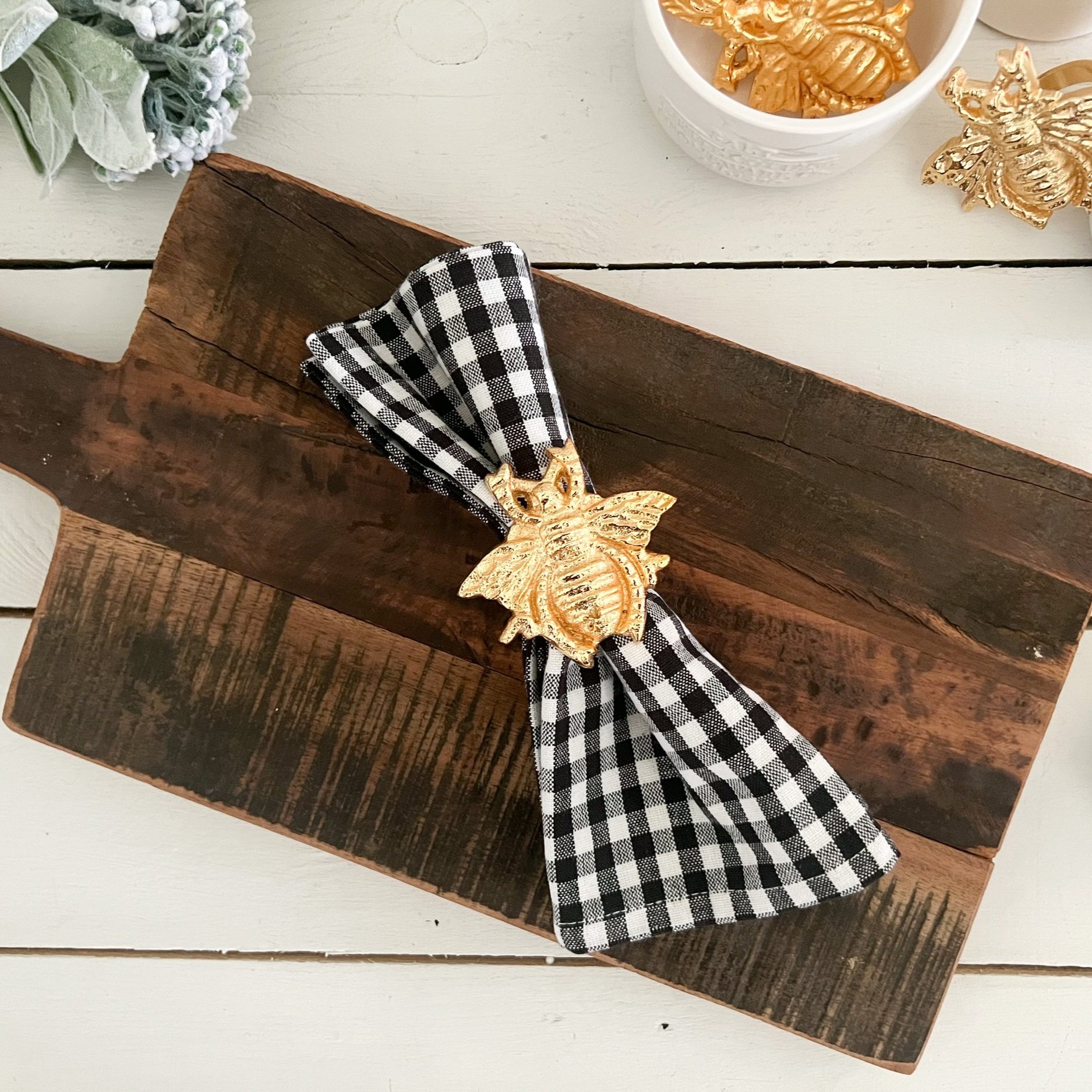 Decorative wooden box with a black and white checkered ribbon, adorned with a gold bee ornament, on a white wooden surface. Partially visible gold bee ornaments, introduction with green foliage and lavender flowers.