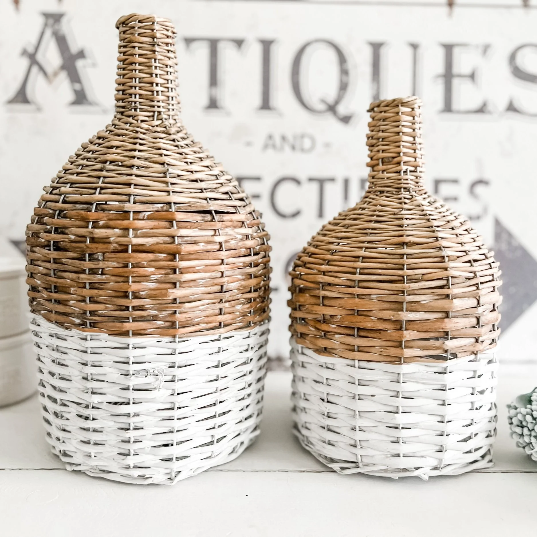 Two decorative woven bottles with white and natural wicker, placed on a white surface with a blurred background of a sign.