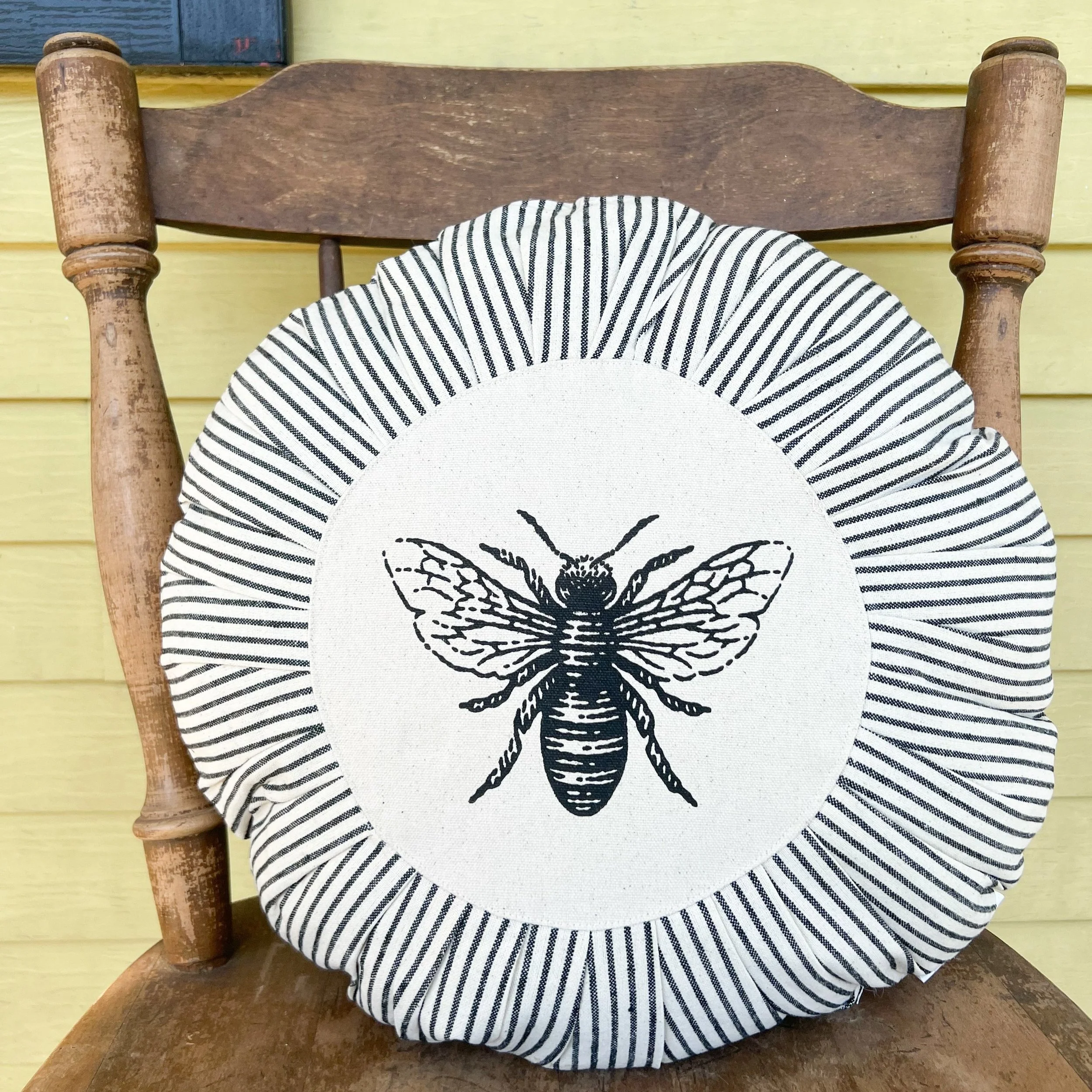 Decorative pillow with a black and white bee illustration, placed on a wooden chair against a yellow wall.