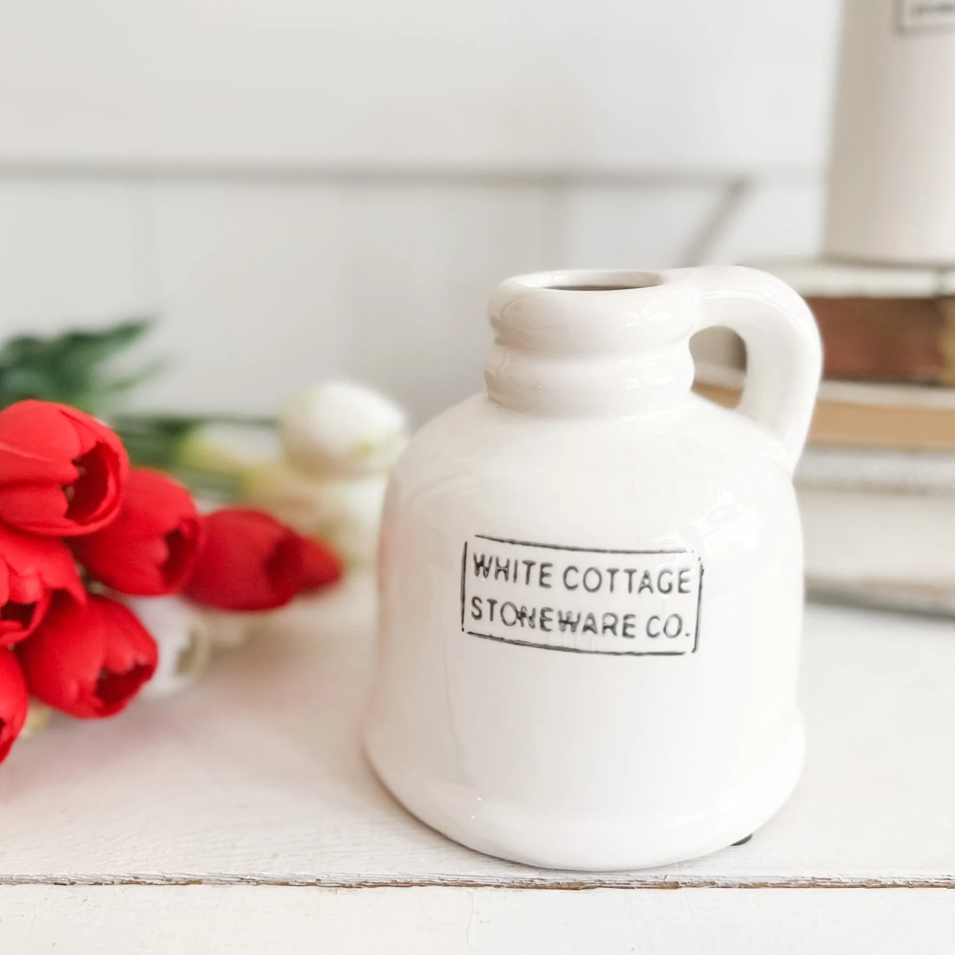A white ceramic jug labeled 'White Cottage Stoneware Co.' with red and white tulips on a white surface, and blurred books and plants in the background.