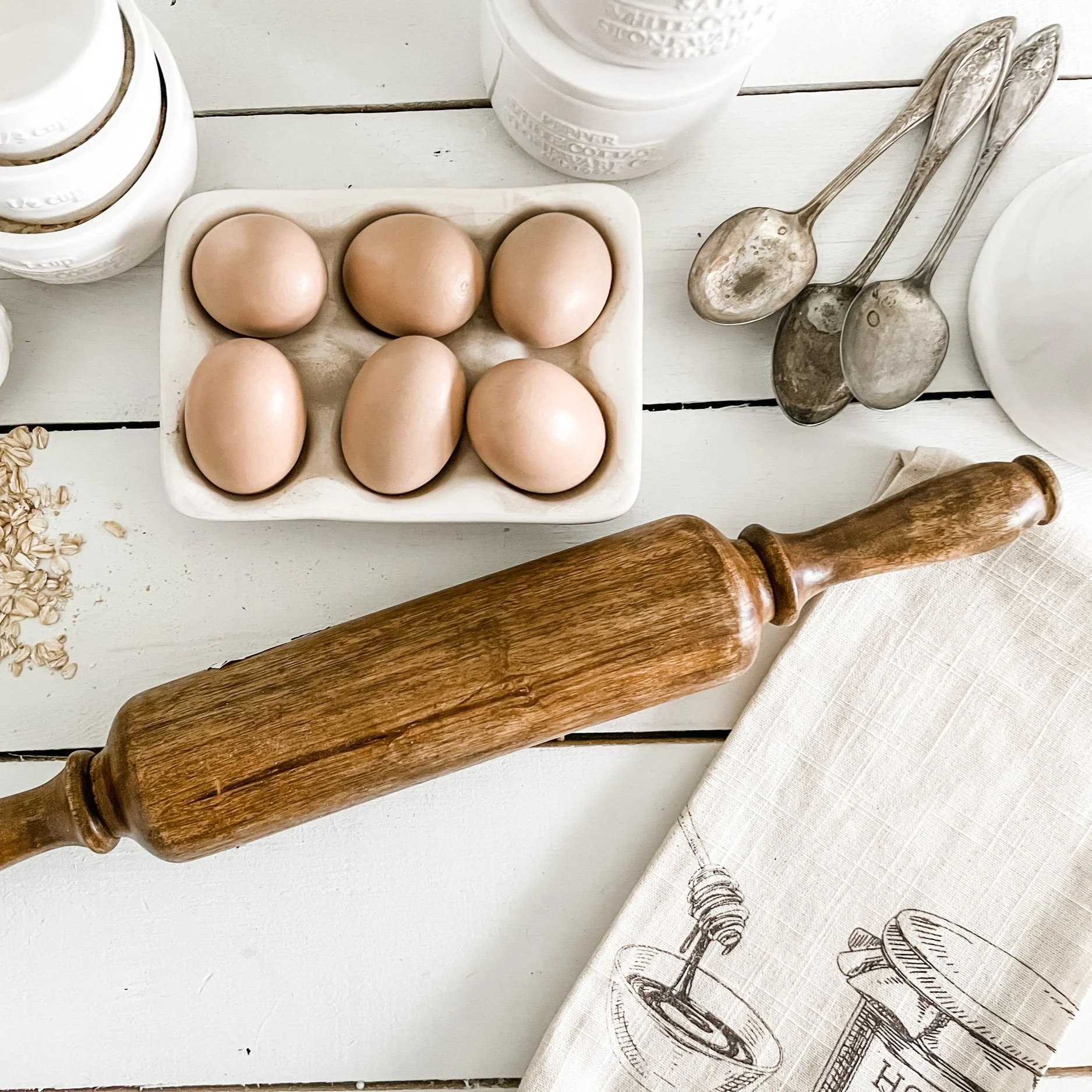 Six brown eggs in a white ceramic tray on a white wooden surface surrounded by metal measuring spoons, a rolling pin, cloth, and jars.