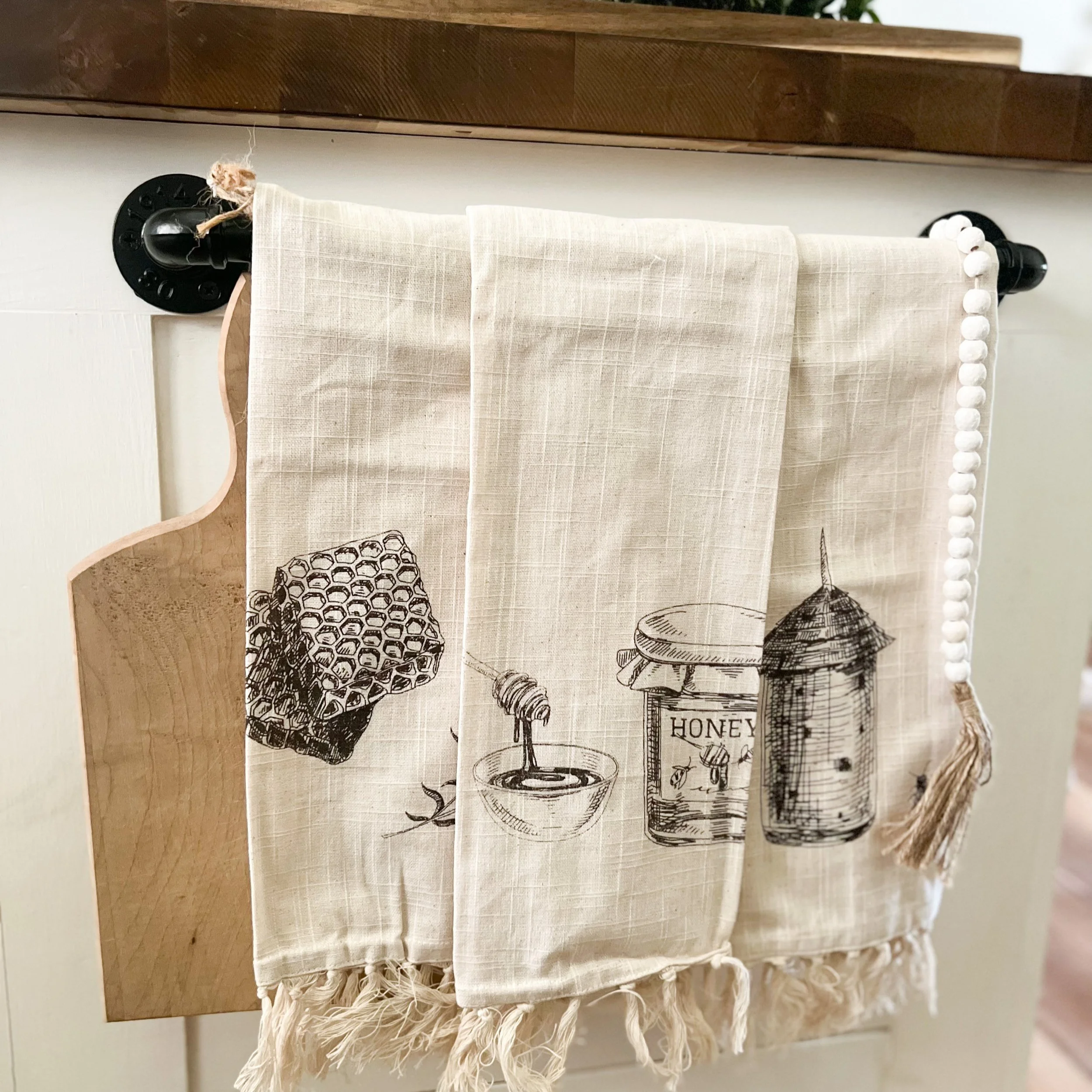 Three beige dish towels with black kitchen-related drawings, hanging on a black towel bar attached to a white wall next to a light wood cutting board.