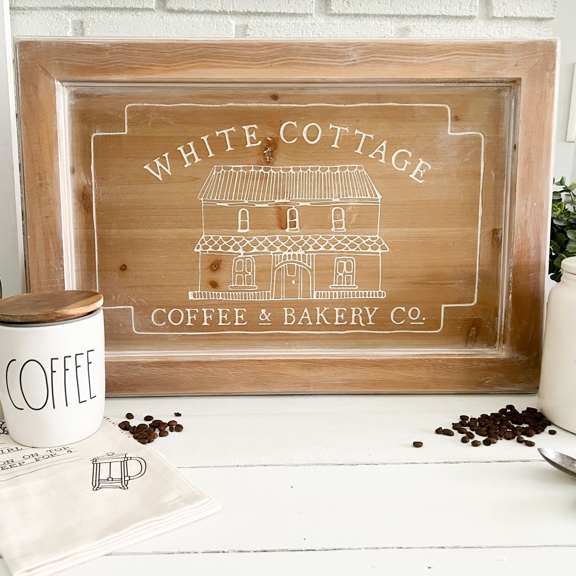 Wooden sign with white writing and illustration of a house that reads 'White Cottage Coffee & Bakery Co.' on a white tabletop, with coffee beans, a white mug with the word 'Coffee' on it, a white bottle, and a spoon nearby.