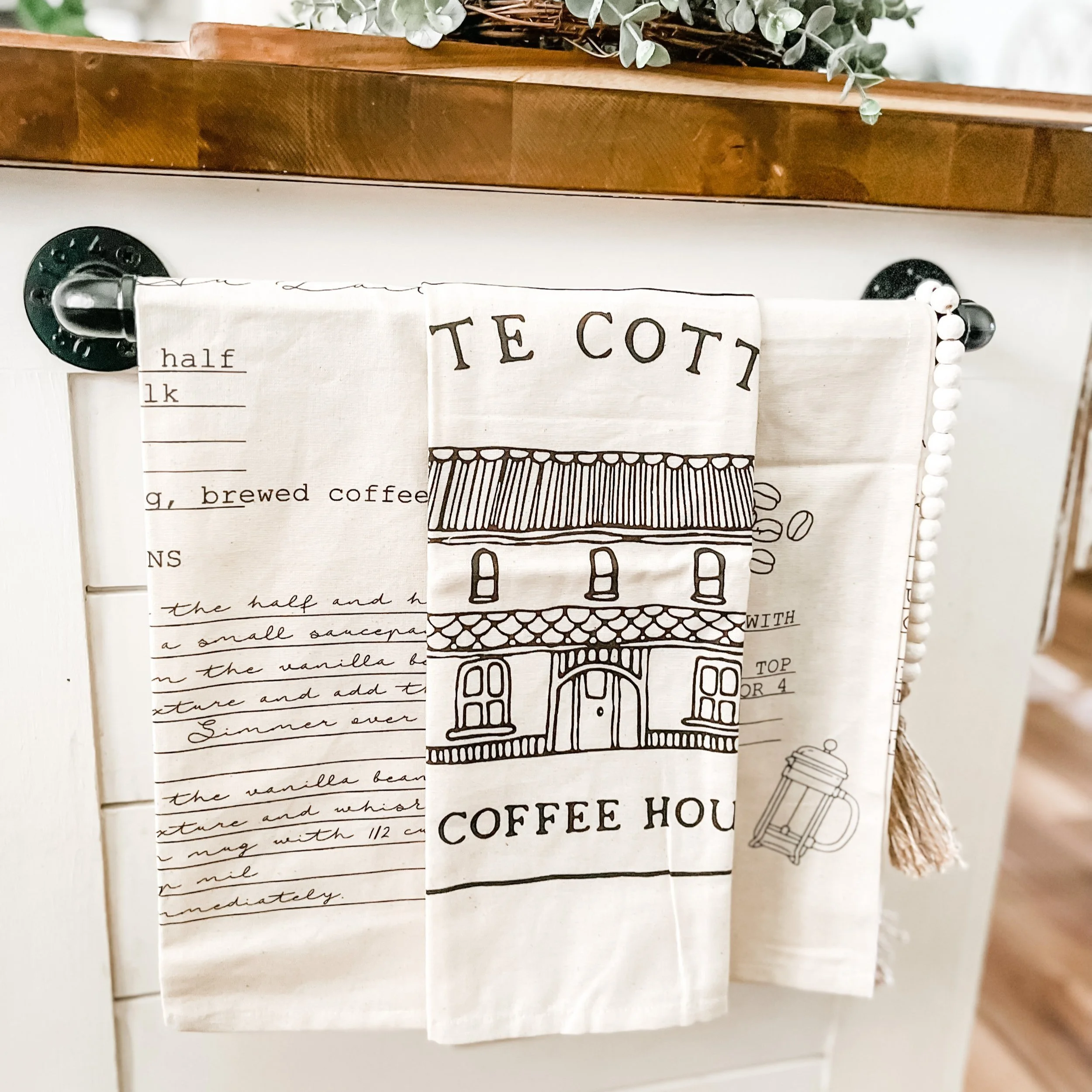 Set of tea towels hanging on a black rod in front of a white cabinet with a wooden top, featuring a building sketch and text about a coffee house.