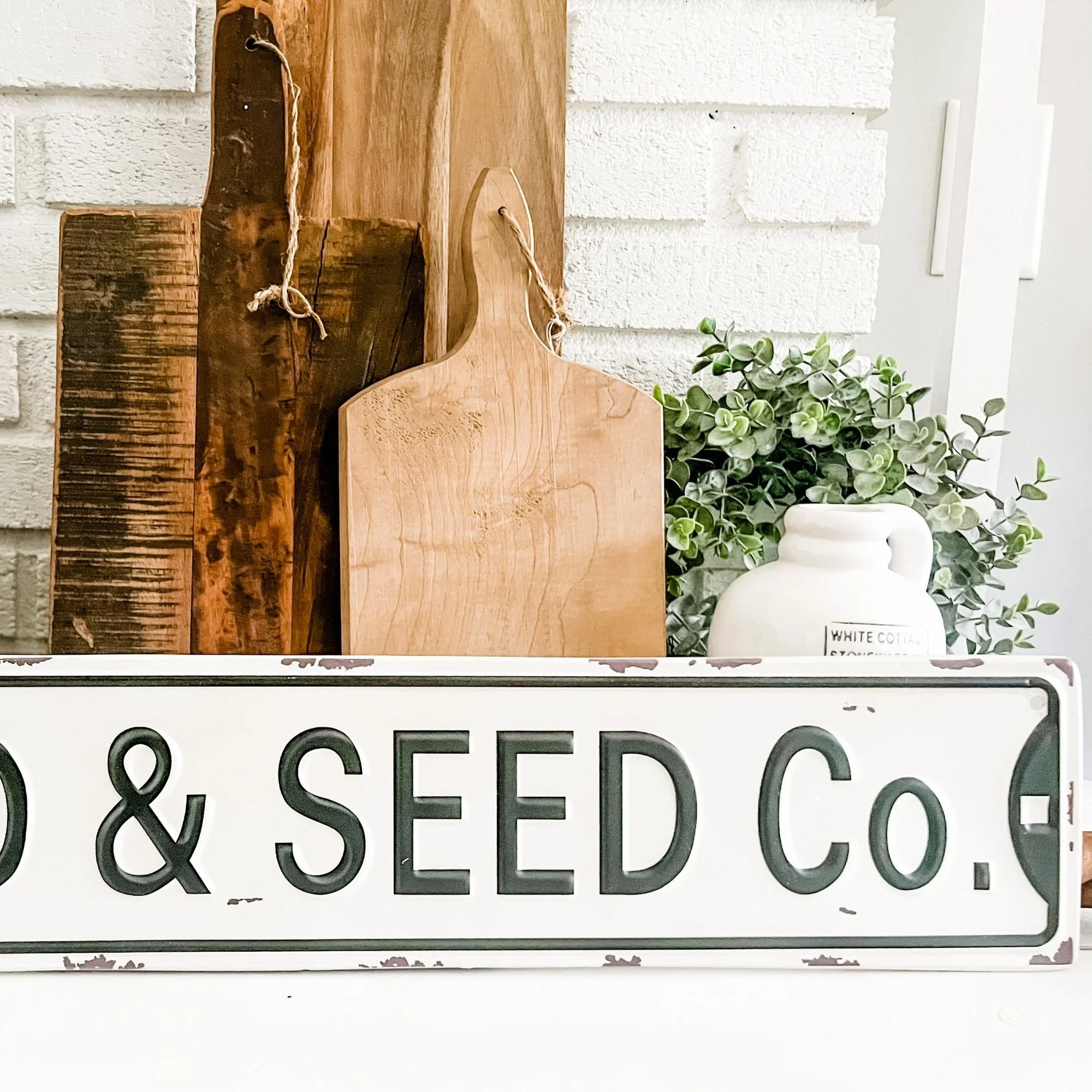 Vintage metal sign with the text '& SEED Co.' placed in front of wooden cutting boards and a white ceramic vase with green leafy plant.