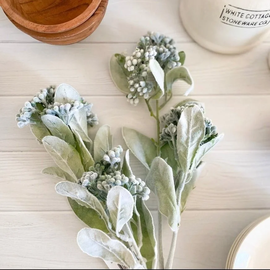 A sprig of artificial eucalyptus with grey-green leaves and white berries, placed on a white wooden surface near a wooden bowl and a white container.