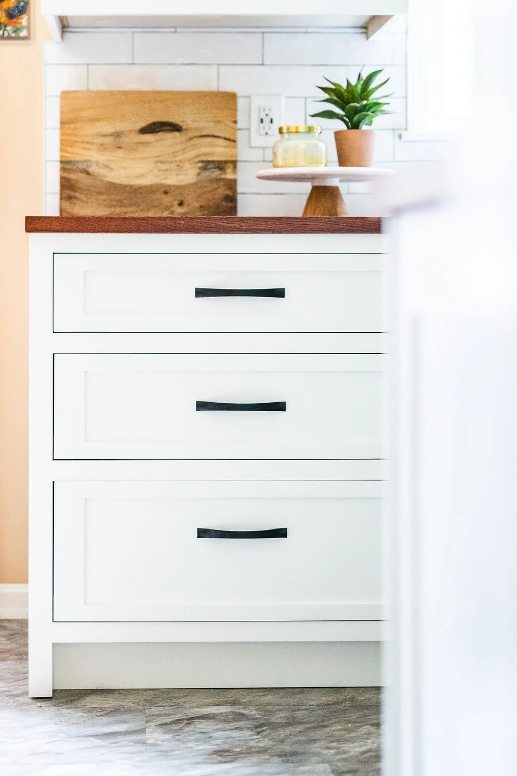 A white kitchen drawer cabinet with three drawers, black handles, and a wooden countertop. Items on top include a cutting board, a potted plant, and a small jar. White tile backsplash and wall are visible in the background.