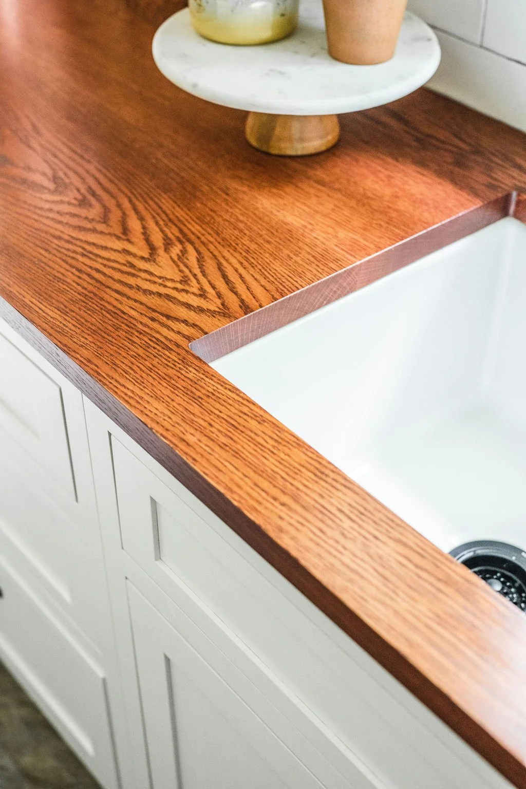 Close-up of a kitchen countertop with a marble cake stand holding a yellow and white cake, and a pink cup on top of the stand. The countertop is made of wood with a visible grain pattern, and there are white cabinets below.