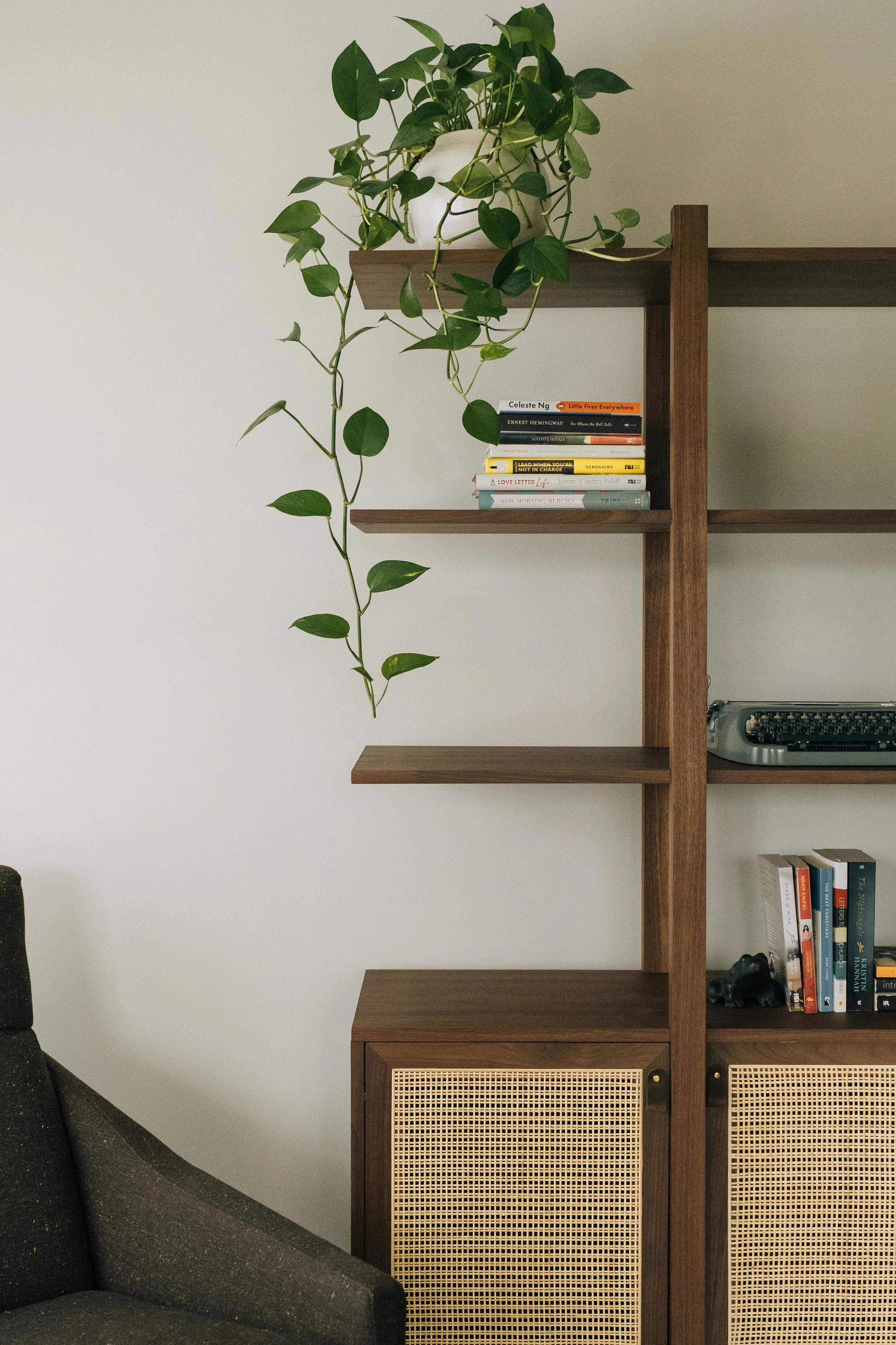 A wooden bookshelf with a potted plant on top, showing green leaves cascading down and several books on the middle shelf, next to a dark gray armchair partially visible on the left.