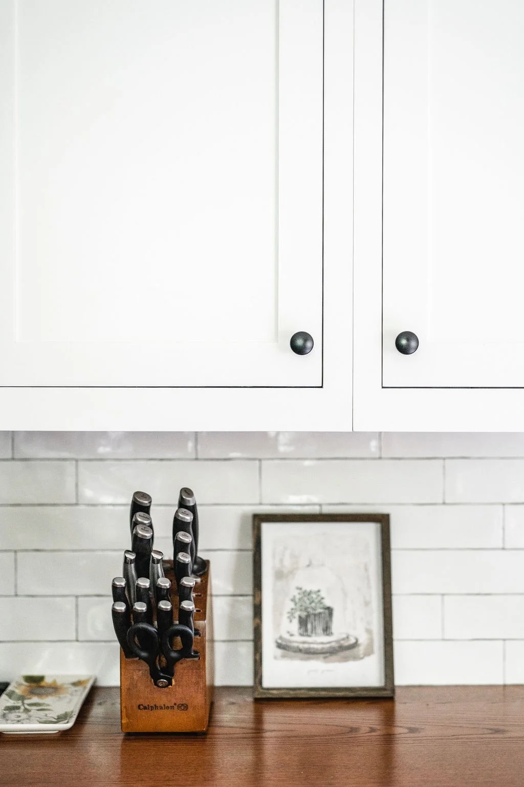 Kitchen with white cabinets, a wooden knife block with black-handled knives, a framed sketch of a potted plant, and a decorative tray on a wooden counter.