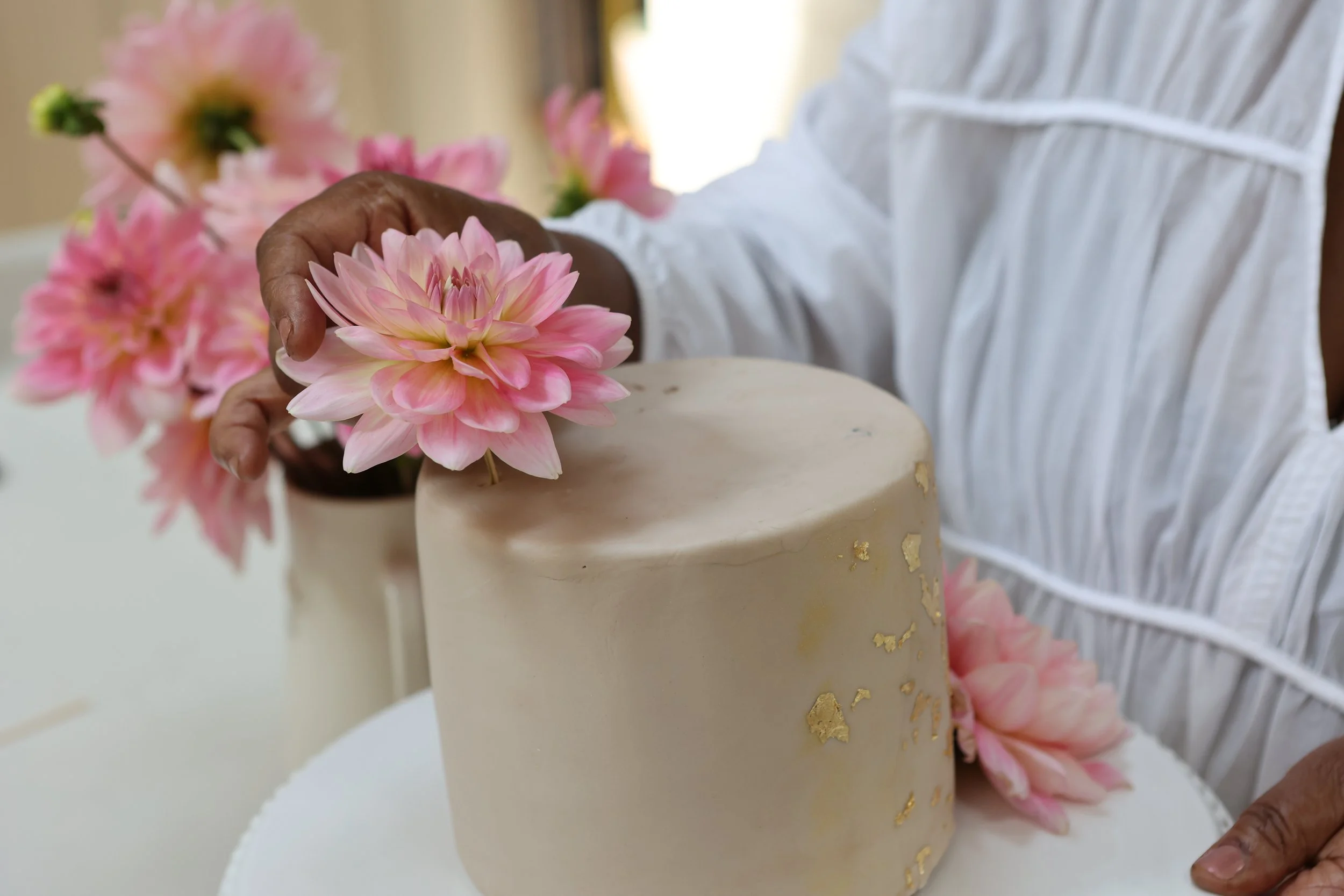 Person placing a pink dahlia flower on a beige cake decorated with gold leaf.
