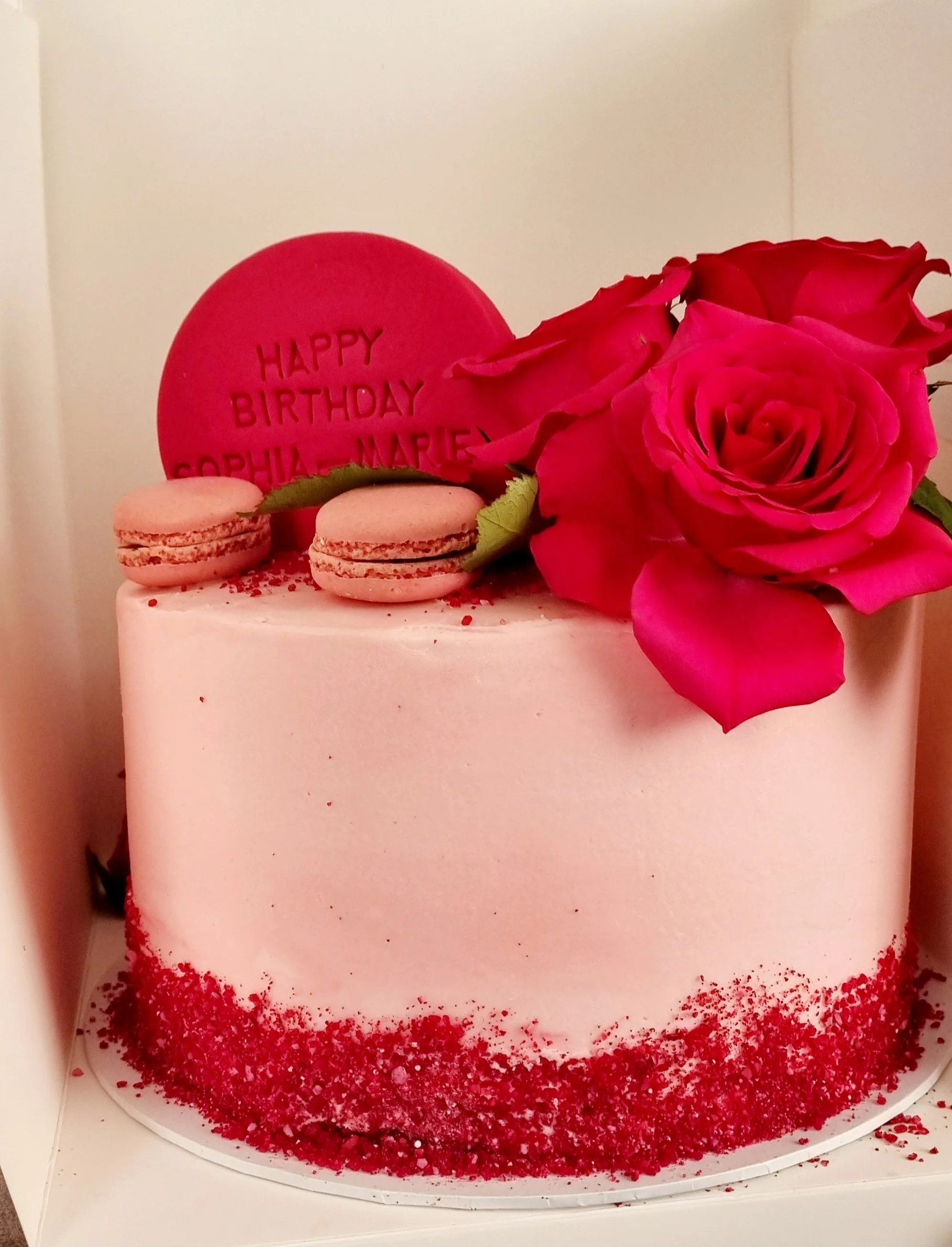 Pink birthday cake with red sprinkles, two pink macarons, a large pink rose, and a red circular cookie with the message 'Happy Birthday Sophia-Marie'