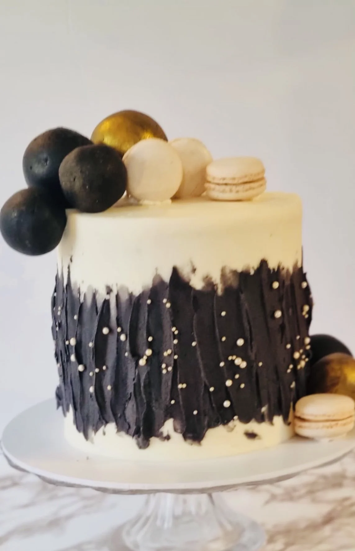 A layered cake decorated with black and white chocolates, macarons, and blackberries on top, with a white and black chocolate design on the side, presented on a glass cake stand.