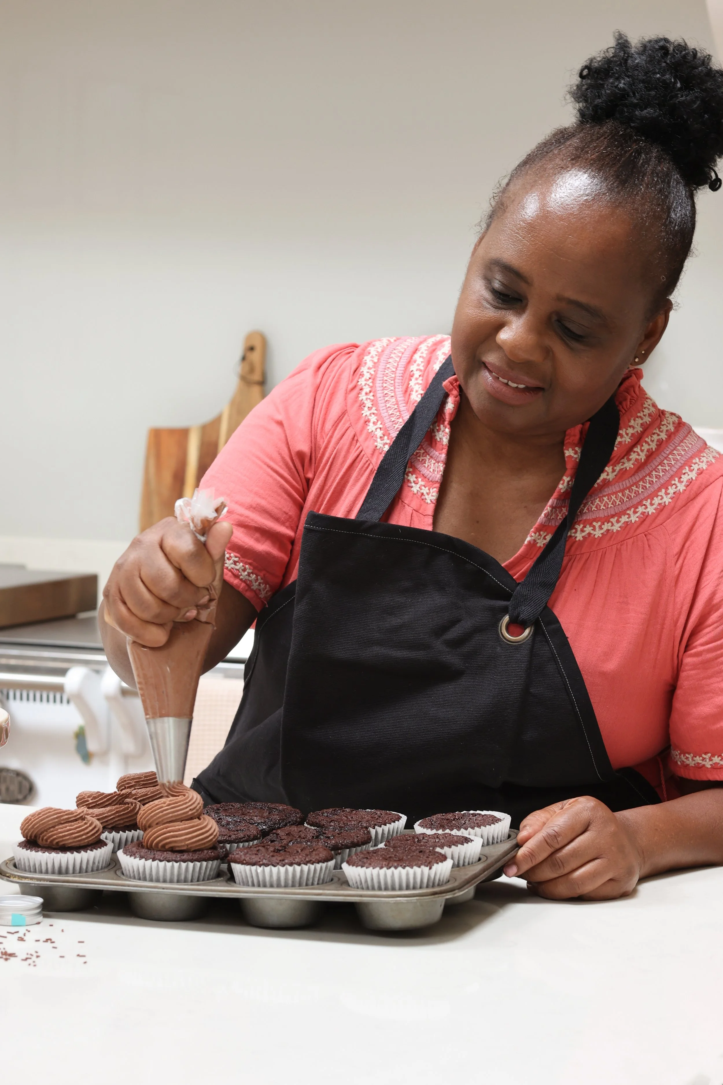 A woman in a pink shirt and black apron decorates chocolate cupcakes with chocolate frosting using a piping bag in a kitchen.