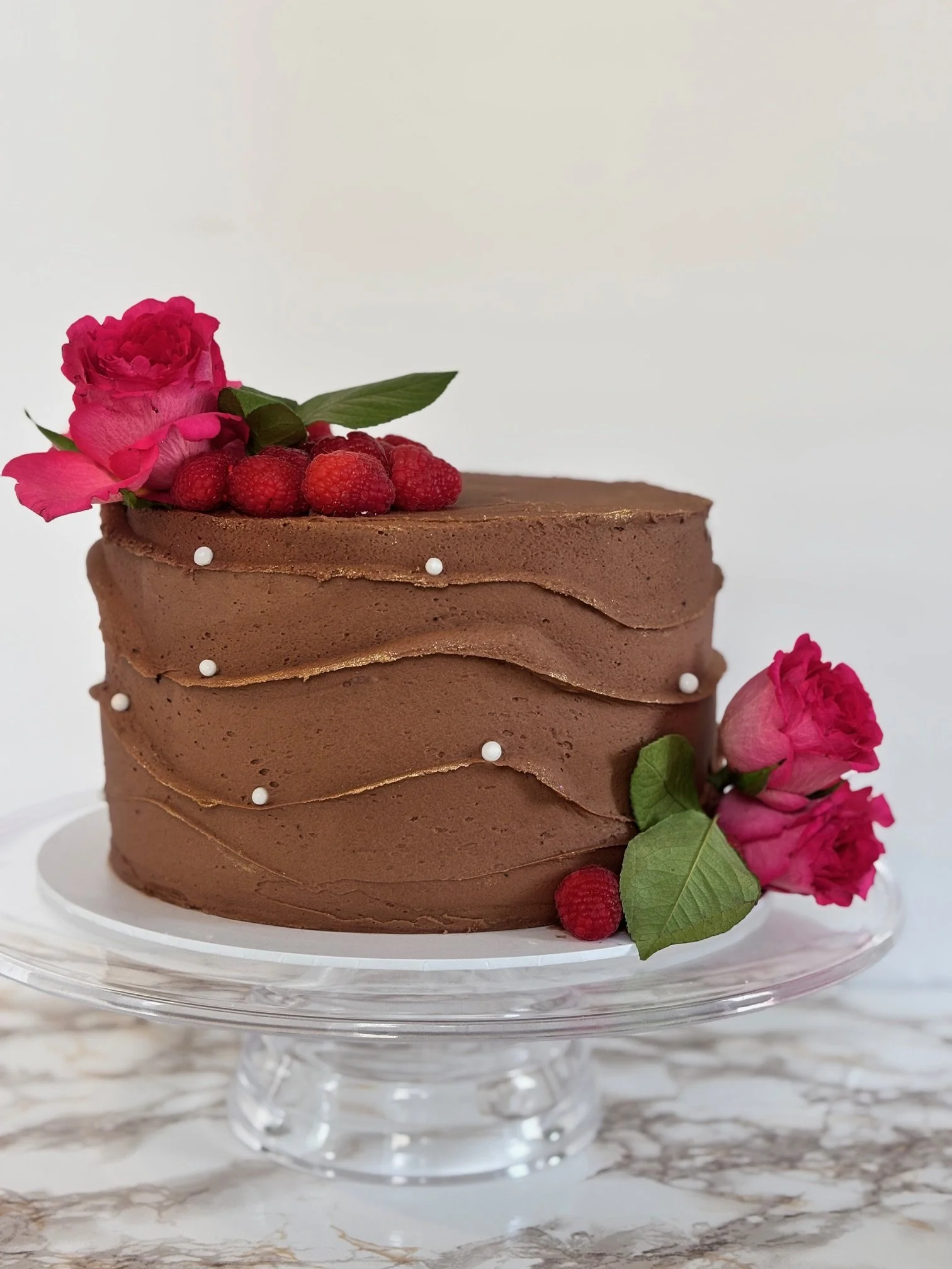 Chocolate layered cake decorated with pink roses, raspberries, green leaves, and small white edible beads on a glass cake stand.