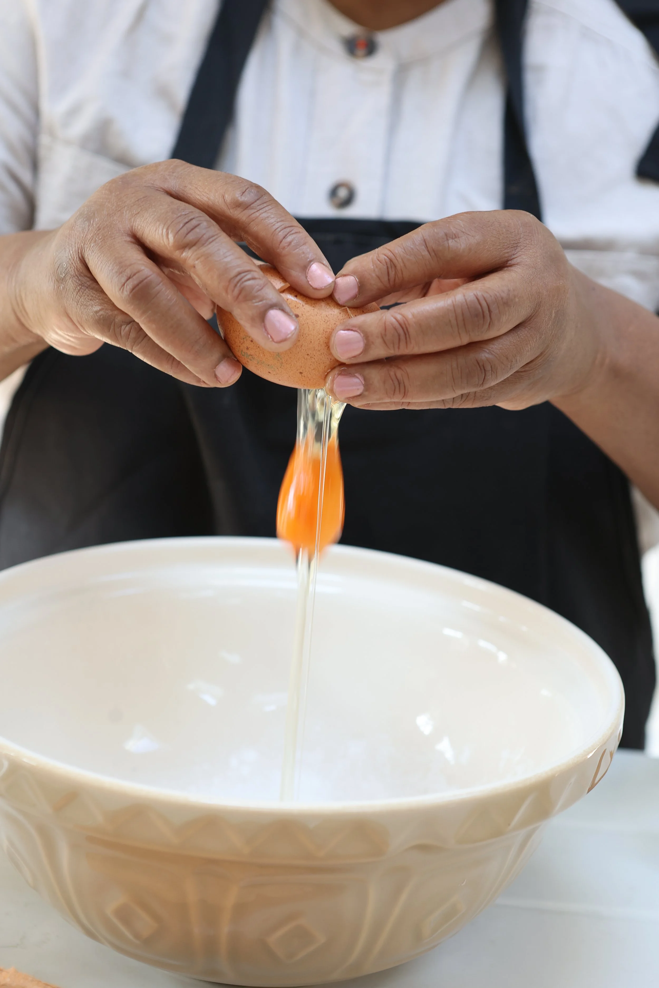 Hands cracking a brown egg over a white mixing bowl, with egg yolk and albumen flowing into the bowl.