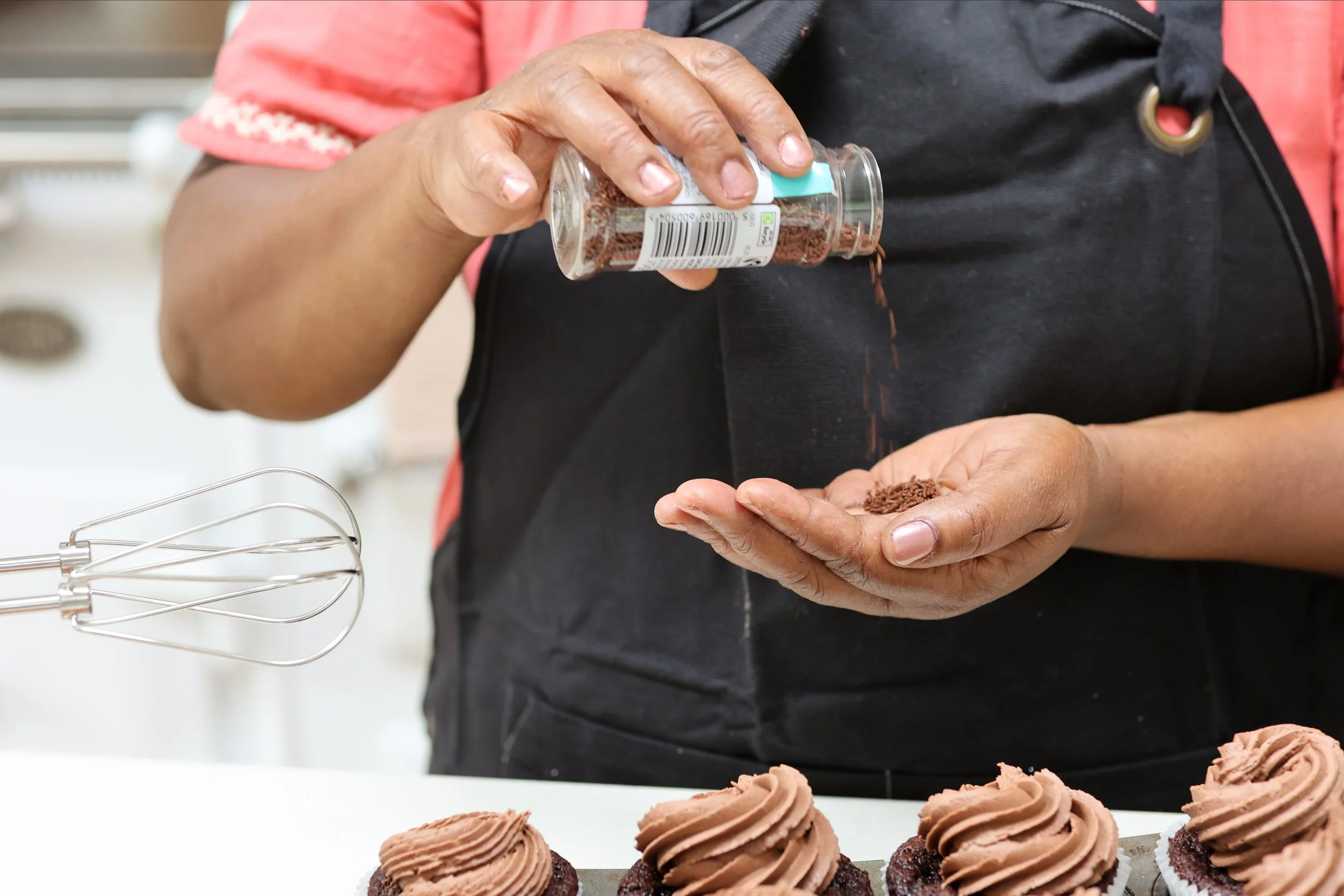 Person sprinkling chocolate sprinkles onto frosted cupcakes in a bakery kitchen.