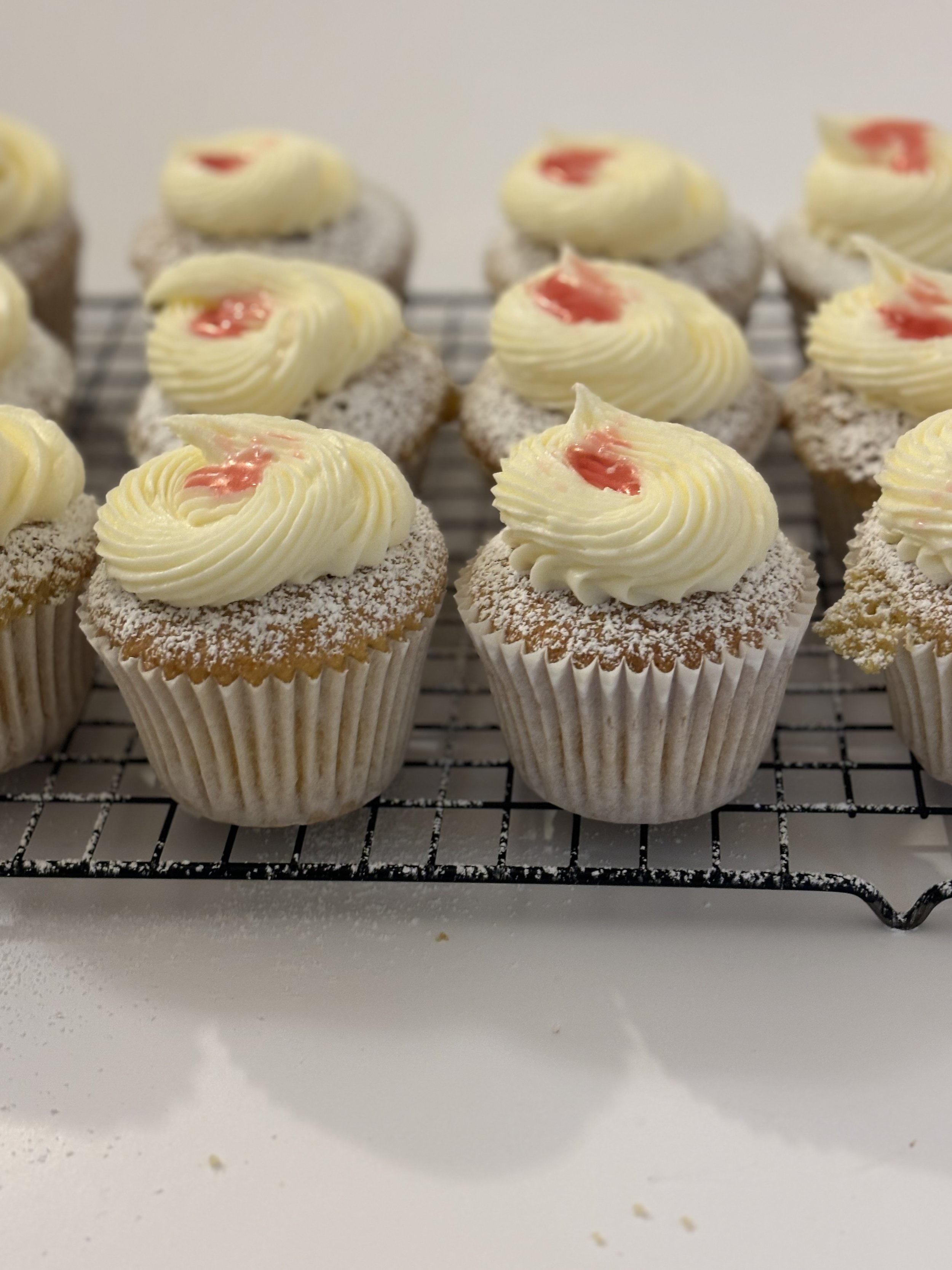 A tray of cupcakes topped with swirls of vanilla frosting and small dollops of pink icing, dusted with powdered sugar.