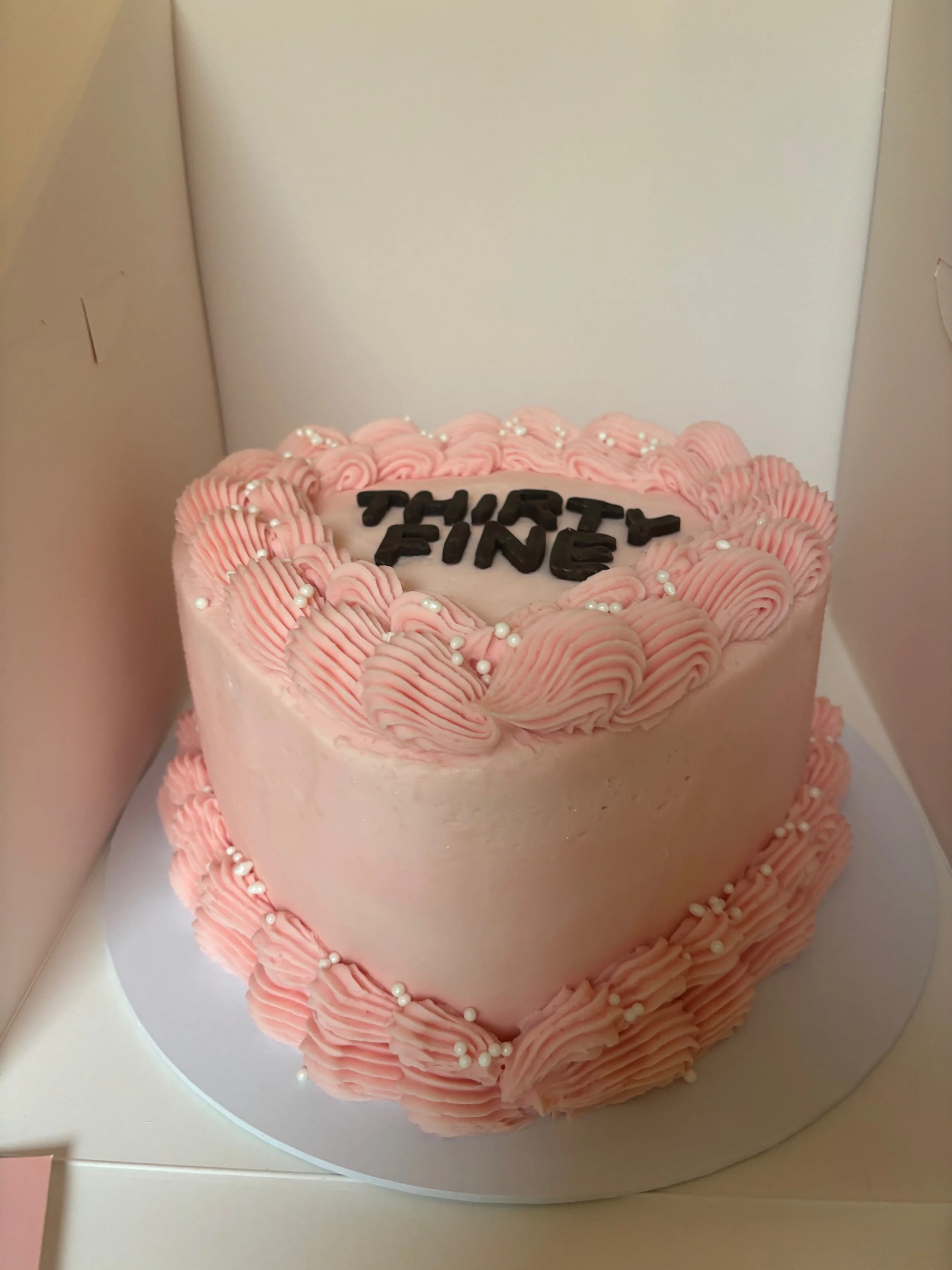 Pink heart-shaped cake with piped pink frosting, white pearl sprinkles, and the words 'THIRTY FIVE' written in black on top.