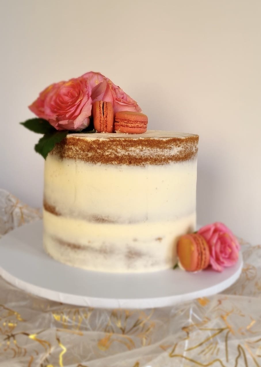 Naked cake decorated with pink roses and orange macarons on top and at the base, on a white cake stand.