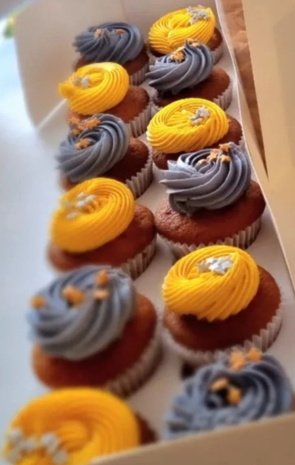 A box of assorted cupcakes with yellow and gray swirled frosting, decorated with small edible toppings.