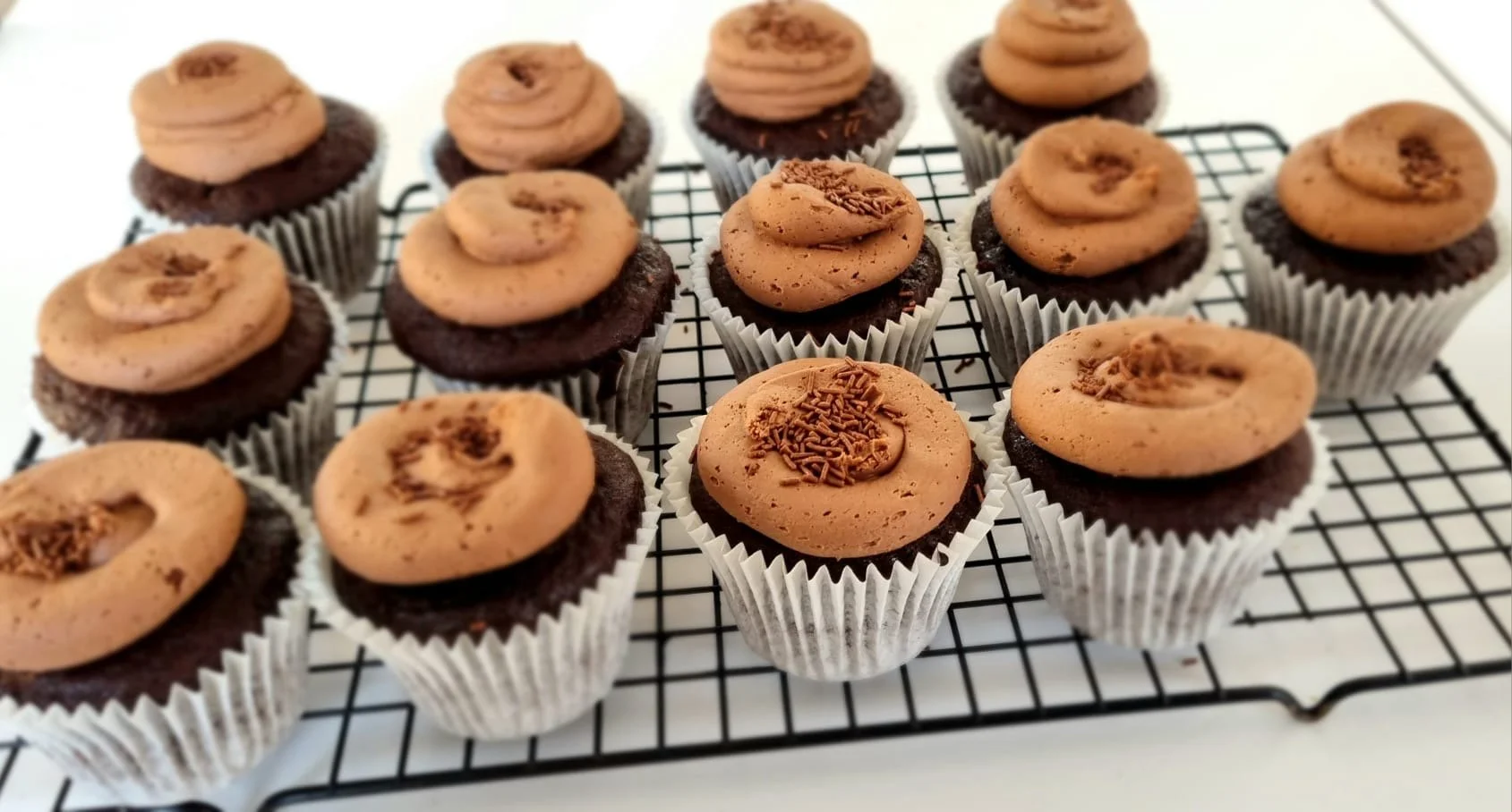 Chocolate cupcakes topped with swirl of chocolate frosting and chocolate sprinkles on a cooling rack.