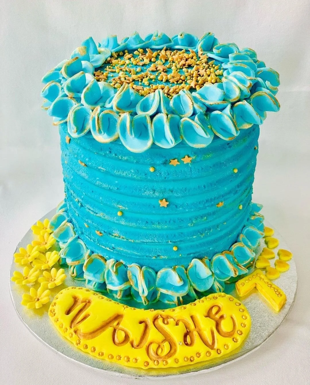 A blue birthday cake decorated with yellow flowers, star and round sugar decorations, and a yellow fondant plaque with a message.
