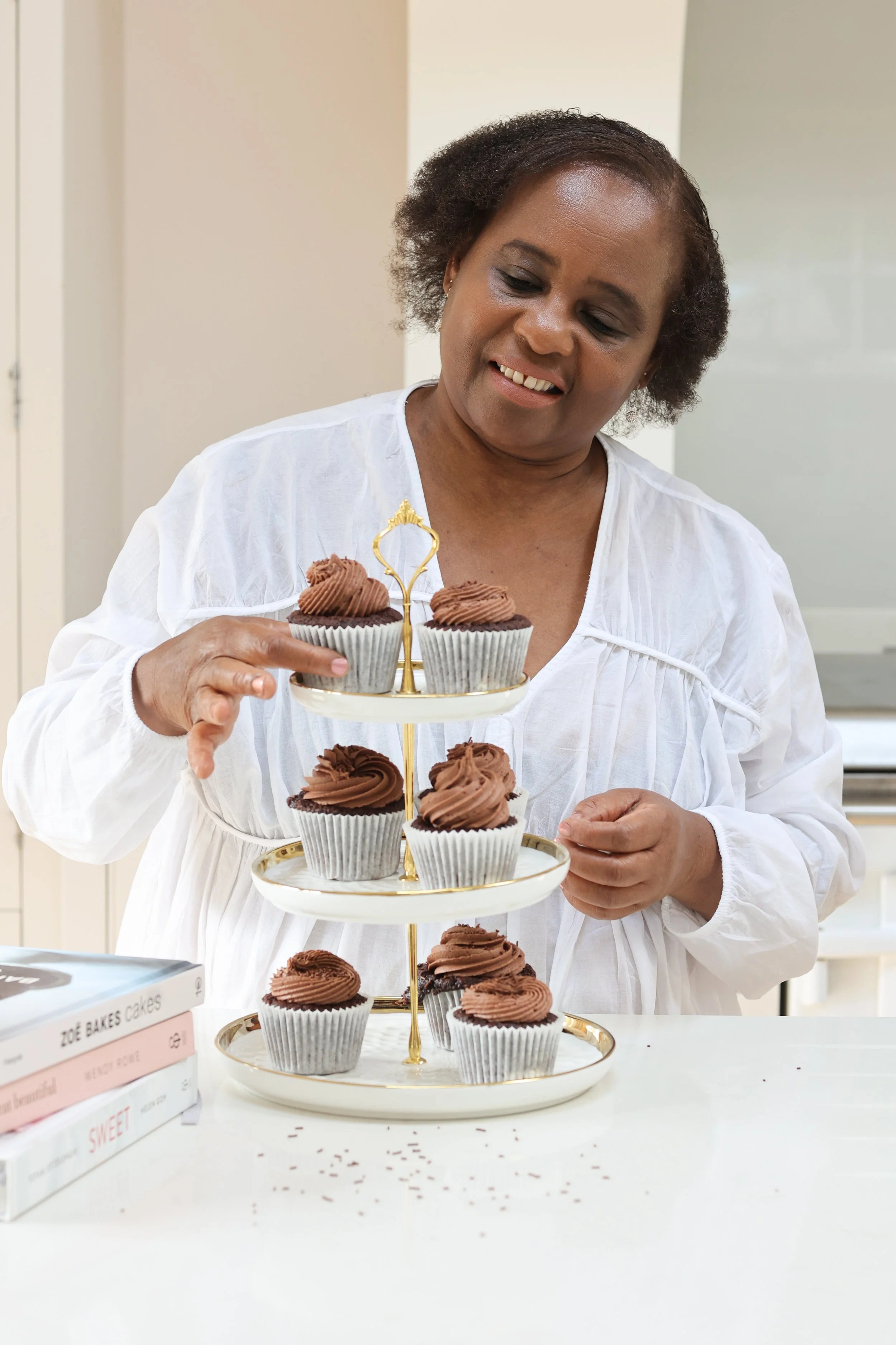 Woman with short curly hair arranging chocolate cupcakes on a two-tiered serving tray in a bright kitchen.