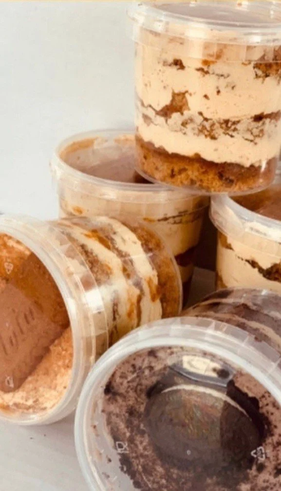 Multiple cups of tiramisu with layers of coffee-soaked ladyfingers, mascarpone cheese, and cocoa powder in clear plastic containers.